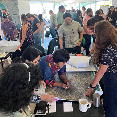 A group of people gather in a bright, open room for a collaborative workshop. Several individuals stand around large printed maps spread across tables, closely examining the materials and writing notes.